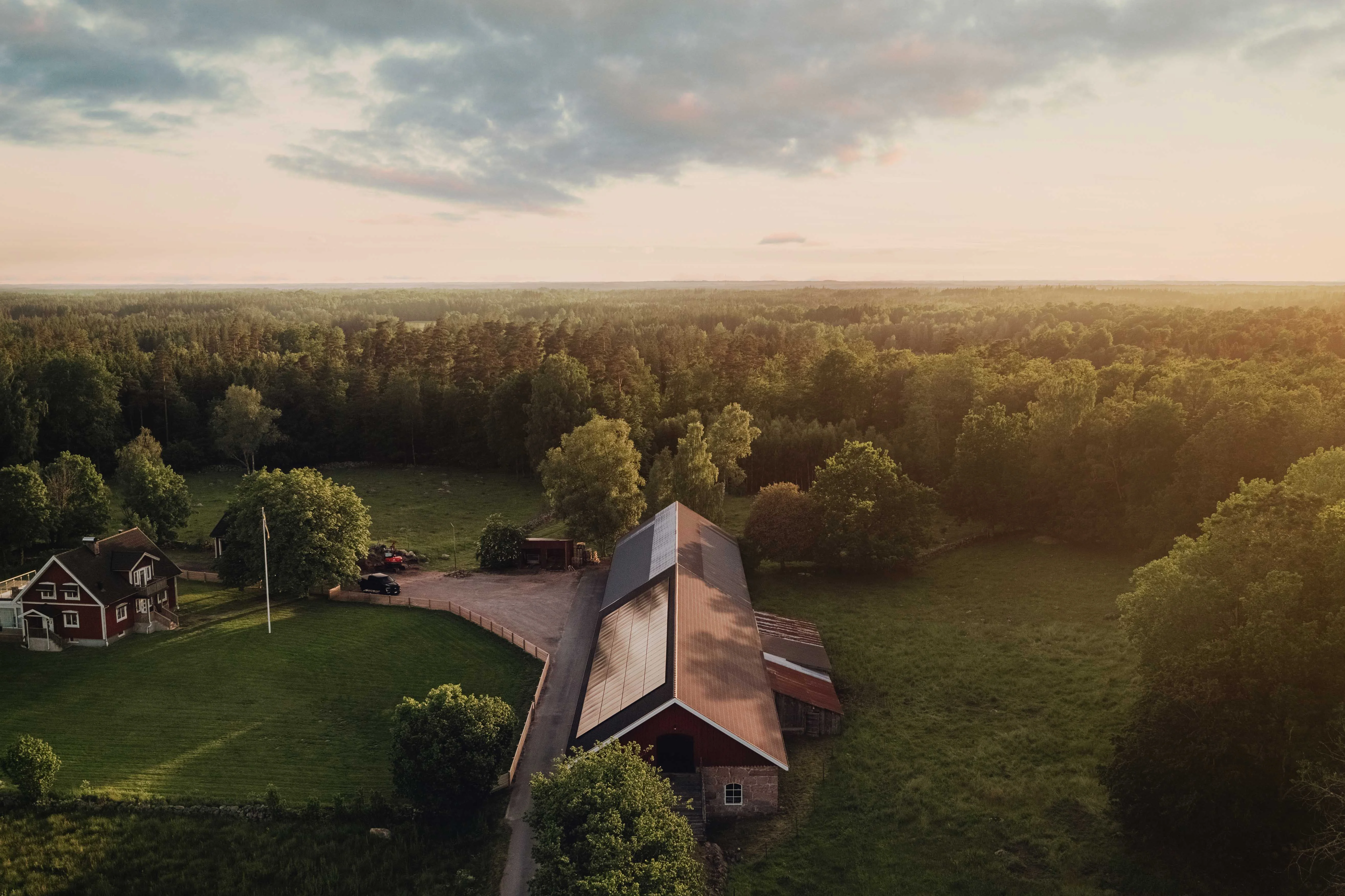 Luftaufnahme eines modernen Holzhauses mit Solaranlage auf dem Dach, eingebettet in eine weitläufige, grüne Wald- und Wiesenlandschaft bei Sonnenuntergang. Das Gebäude steht inmitten der Natur, umgeben von Bäumen und Nebenhäusern.